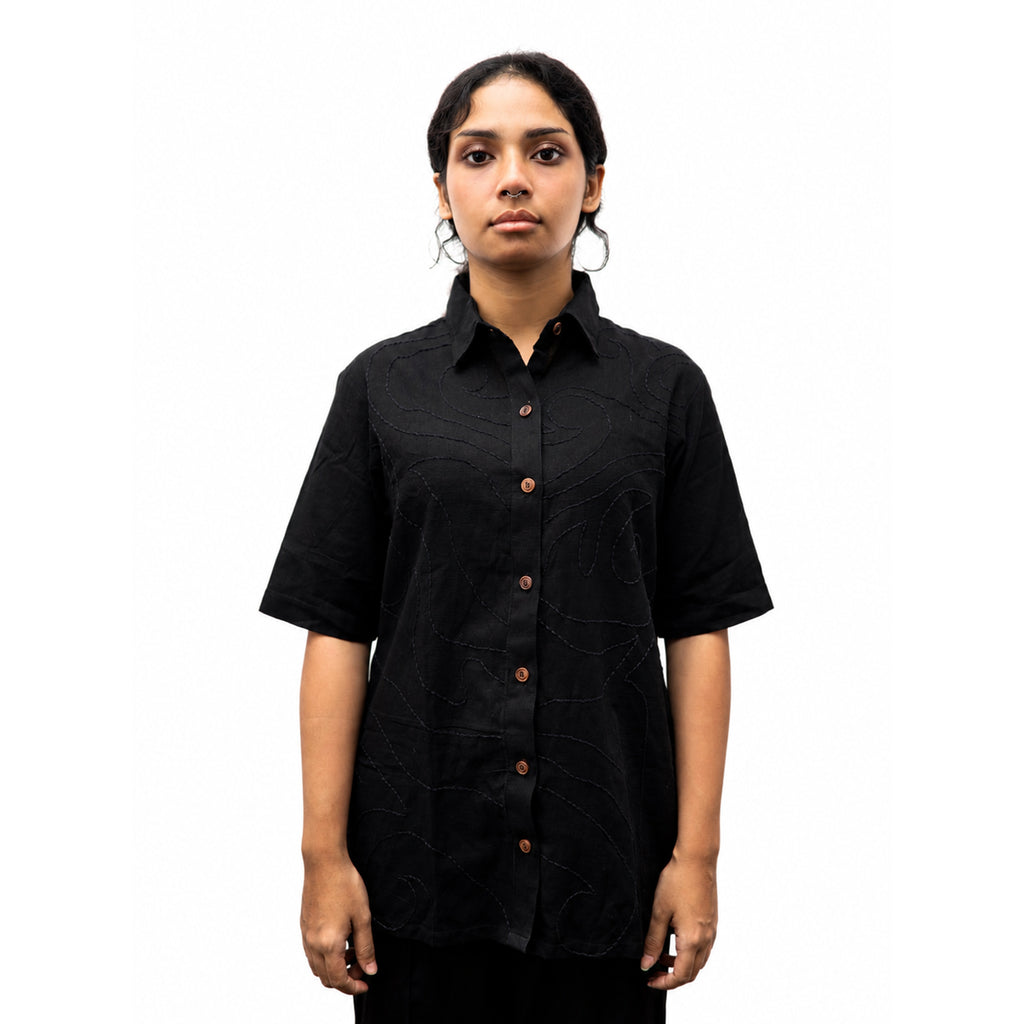 A woman modeling a black short-sleeve button-up shirt with a subtle tonal abstract pattern and brown buttons, set against a plain white background.