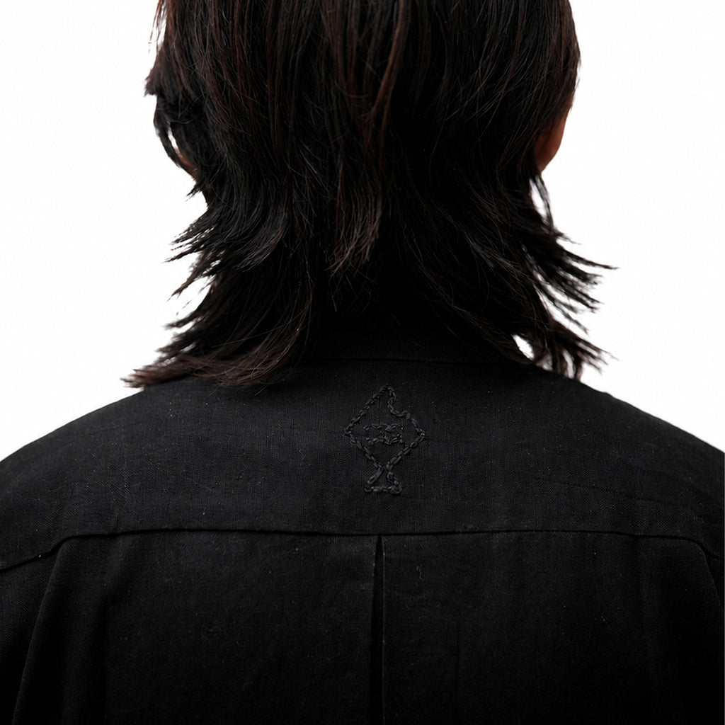 Back view of a person with shoulder-length dark hair wearing a black collared shirt or jacket, showing a small tonal embroidered emblem centered on the upper back against a white background.